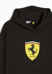 Svart huvtröja med en framträdande gul Ferrari-logotyp med en sprattlande häst, inramad av röda och gröna linjer, som visar en sportig design.