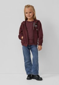 Burgunderfarbene Zip-Up-Hoodie mit gerippten Bündchen, über einem weinroten Langarmshirt und blauen Jeans getragen, ergänzt durch schwarze Schuhe.