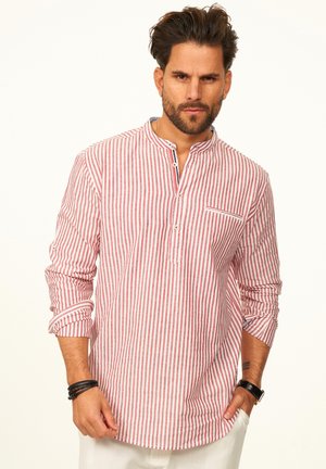 Mann mit Bart, trägt ein rot-weiß gestreiftes Hemd mit Stehkragen und hochgekrempelten Ärmeln sowie eine weiße Hose.