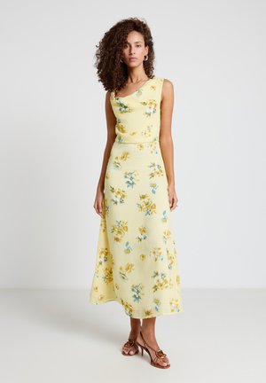 Femme portant une robe midi jaune fleurie sans manches avec un décolleté asymétrique et des sandales marron à talons, debout devant un fond uni.