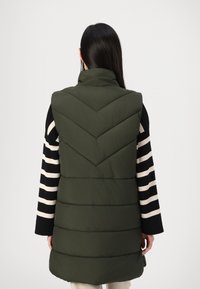 Grønn polstret vest med høy krage, med en chevron-quiltet design og sidelufter, sammen med en svart-hvit stripete langermet topp.