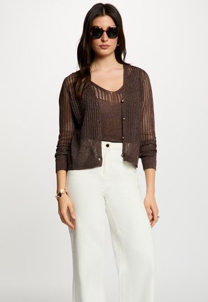 Donna che indossa un cardigan in maglia marrone scuro trasparente e top, con pantaloni bianchi a vita alta a gamba larga e occhiali da sole scuri oversize, in piedi davanti a uno sfondo semplice.
