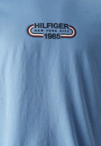 Jasnoniebieska tkanina z napisem "HILFIGER NEW YORK CITY 1985" oraz czerwonymi, białymi i niebieskimi zakrzywionymi paskami nad i pod napisem.