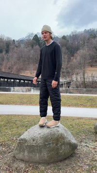 Sort langærmet skjorte og puffede sorte bukser, stående på en sten. Beige sko og en lys hue, med et bjerglandskab i baggrunden.