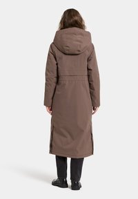 Bruine lange jas met een capuchon, gemaakt van gladde stof, met zijsplitten en gestikte details, gedragen over zwarte broek en laarzen.