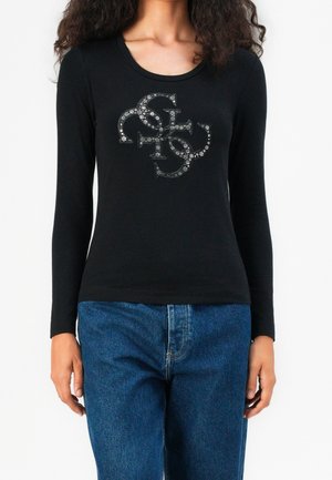 Kvinde iført sort langærmet bluse med pyntet blomsterlogo design og blå jeans, vist fra hals til hofter på hvid baggrund.