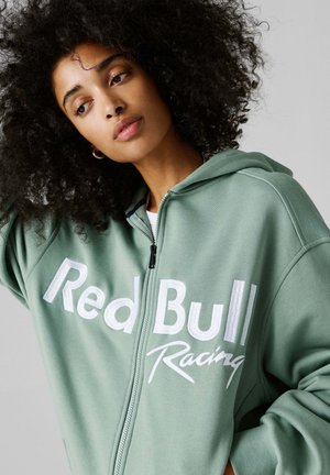 Moteris su garbanotais plaukais, dėvinti žalią su užtrauktuku striukę su užrašu „Red Bull Racing“, žiūri šiek tiek į šoną priešais paprastą foną.