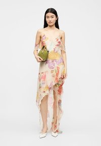 Femme portant une robe midi fleurie en tissu transparent avec un ourlet asymétrique à volants, tenant un petit sac à main vert olive et chaussée de chaussures blanches à bout pointu.