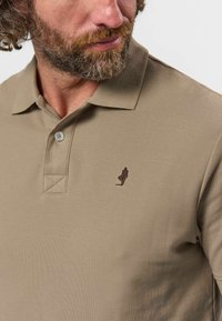 Homme avec barbe portant un polo beige avec un petit logo figurine brodé foncé sur la poitrine, regardant sur le côté.