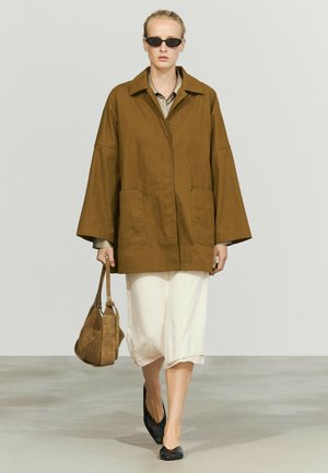 Frau, die eine übergroße braune Jacke, einen weißen Rock, schwarze flache Schuhe, eine dunkle Sonnenbrille trägt und eine braune Wildledertasche bei sich hat.