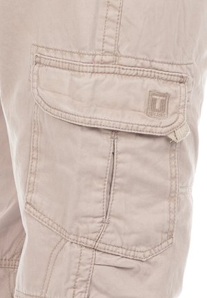 Beige cargo pants with multiple pockets, made from soft fabric. Visible stitching details and a small logo patch on the front pocket.