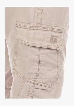 Beżowe spodnie cargo z wieloma kieszeniami, wykonane z miękkiego materiału. Widoczne detale szycia oraz mała naszywka z logo na przedniej kieszeni.