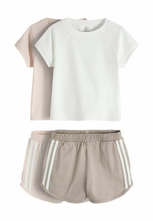 Ung pige med langt brunt hår står mod en lys væg iført en hvid t-shirt, beige shorts med striber, brune sokker og hvide sneakers.