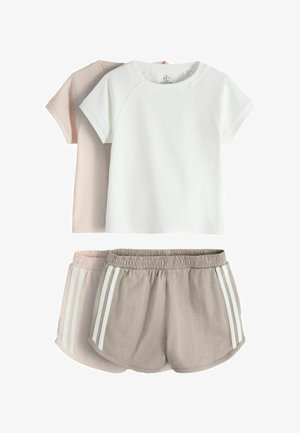 Ung pige med langt brunt hår står mod en lys væg iført en hvid t-shirt, beige shorts med striber, brune sokker og hvide sneakers.
