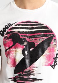 Vit t-shirt med en svart surfbräda grafiskt motiv med en siluett av en surfare mot en rosa och svart cirkulär bakgrund. Texten ovanför säger "Ooooh..."