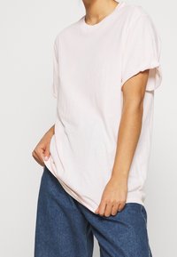 Jasnoróżowa, oversize koszulka z krótkimi, podwijanymi rękawami, wykonana z miękkiego bawełnianego materiału. W zestawie ze luźnymi niebieskimi dżinsami.