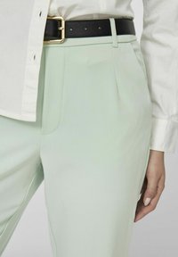 Pantalons sur mesure vert clair avec plis avant, poches latérales et une ceinture noire avec une boucle en or, associés à une chemise blanche à boutons.