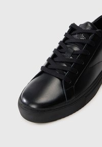 Black leather sneaker with a smooth finish, round toe, flat black laces, and a textured rubber sole. Features minimal stitching details.