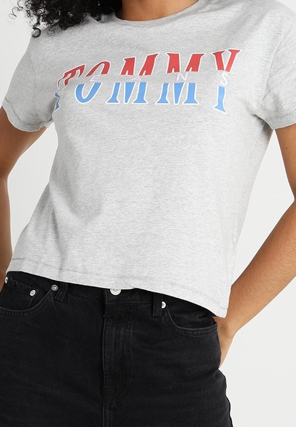 Camiseta de algodón gris con el texto "TOMMY" en rojo y azul, con un diseño corto y mangas cortas, combinada con jeans negros.