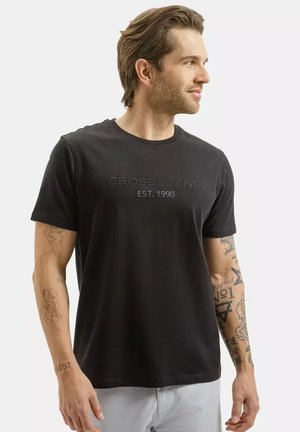 Mężczyzna z tatuażami na obu ramionach, ubrany w czarną koszulkę z napisem „CROSS JEANS EST. 1990” i jasne spodnie, patrzący w prawo.