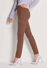Frau in braunen Cargo-Hosen mit Seitentaschen, einem lilafarbenen Pullover und lilafarbenen High-Heels, steht vor einem schlichten hellen Hintergrund.
