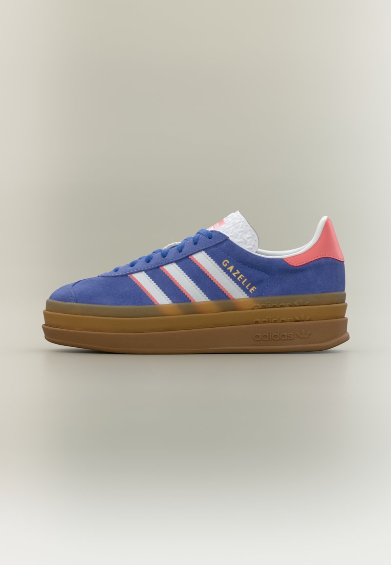 Niebieski sneaker Adidas Gazelle z białymi paskami, różowym zapiętkiem i grubą, brązową, warstwową podeszwą na jasnym, neutralnym tle.
