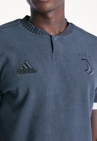 Granatowa koszulka polo z teksturowanej tkaniny, z prążkowanym kołnierzykiem i trzema guzikami. Wyszywane logo Adidas i Juventus w kolorze czarnym.