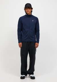 Marine blauwe ritsjack met hoge kraag, gecombineerd met zwarte cargobroeken. Geribbelde stof, zijzakken en logo-accent op het jack.