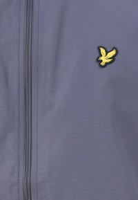 Tissu bleu avec une fermeture éclair verticale et un petit emblème d'oiseau jaune sur le côté droit.