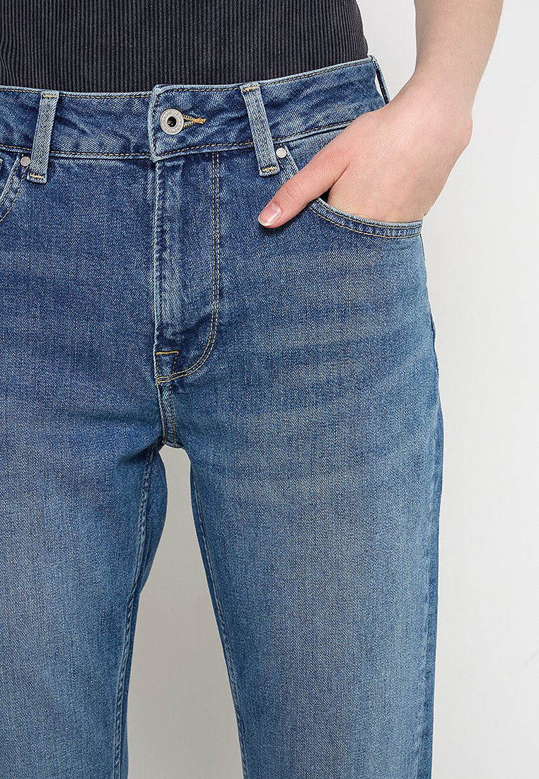Person, der eine blaue Denim-Jeans trägt, mit einer Hand teilweise in der vorderen Tasche vor einem einfarbigen Hintergrund.