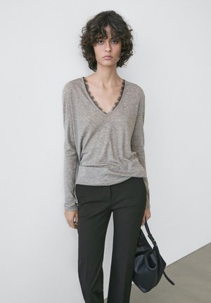 Žena s kudrnatými vlasy, oblečená v šedém tričku s výstřihem do V a krajkovým lemováním, a černých kalhotách, držící černou kabelku na pozadí jednobarevné zdi.