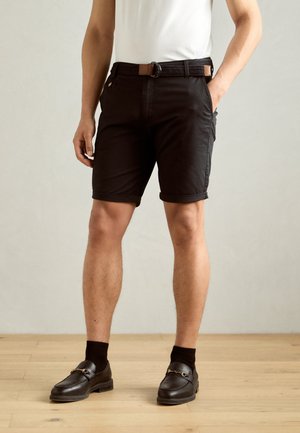 Sorte shorts med oppbrettede kanter, laget av solid stoff. Brukt med en hvit skjorte og et flettet belte. Sorte loafers og sokker fullfører antrekket.