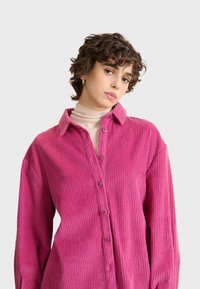 Camisa de pana rosa con cierre de botones, cuello puntiagudo y textura acanalada, con mangas largas y botones visibles.