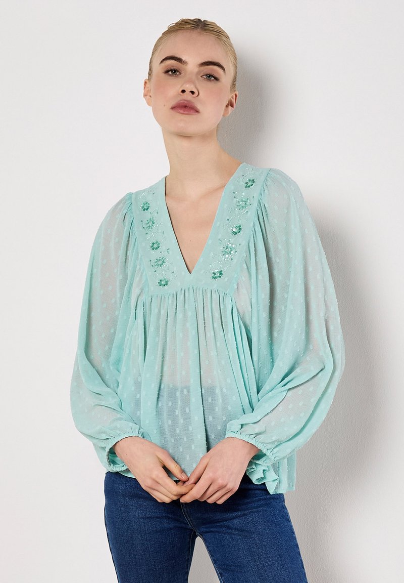 Helles teal-blaues Blusentop aus transparenter Stoff, mit V-Ausschnitt, langen Puffärmel, gepunkteter Textur und floralen Stickereien entlang des Ausschnitts.