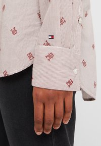 Gros plan sur une main à côté d'un poignet de chemise beige rayé avec des motifs rouges brodés et un petit logo drapeau rouge, blanc et bleu.