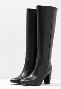 Botas de cuero negro hasta la rodilla con un acabado brillante, punta cuadrada y tacón grueso. Diseño sin costuras con acentos mínimos.