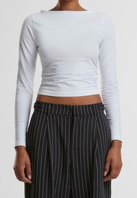 Weißes kurz geschnittenes Oberteil mit langen Ärmeln und weitem Ausschnitt, kombiniert mit schwarzen hochtaillierten Hosen mit Nadelstreifen, die eine glatte Stofftextur zeigen.