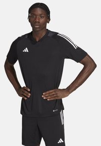 Zwarte sporttrui met korte mouwen, mesh structuur, ronde hals en witte drie-streep accenten op de schouders. Draagt bijpassende zwarte shorts.