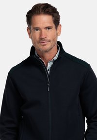 Zwarte zip-up jack met een hoge kraag, gemaakt van gladde stof. Heeft groene schouderaccenten en een ritssluiting, gedragen over een geblokt shirt.