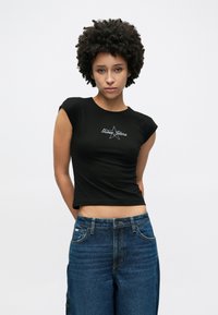 Zwart nauwsluitend T-shirt met korte mouwen en een ronde hals, met het "Guess Jeans"-logo in witte letters en een sterontwerp. Gecombineerd met blauwe jeans.