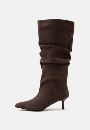 Botte ample en daim brun foncé mi-mollet avec un bout pointu et un talon bas et fin sur un fond clair uni.