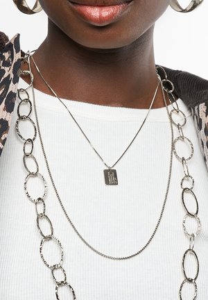 Colliers superposés en ton argent avec un pendentif rectangulaire plat et une chaîne à maillons ovales texturés, portés sur un haut blanc à côtes.
