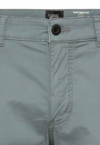 Nahaufnahme des Hosenbunds einer hellgrauen Hose mit schwarzem Knopf, Gürtelschlaufen und sichtbarem, eingenähtem Reißverschlussdetail.