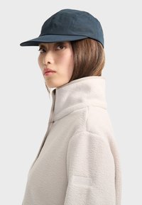 Gorra de marina con ala ancha y tela texturizada, combinada con una chaqueta de forro polar color beige claro con cuello alto y logo en la manga.