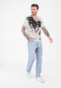 Man met volarmp Tattoos die een wit T-shirt met grafisch schedel draagt, lichtblauwe jeans en witte sneakers, lopend tegen een witte achtergrond.