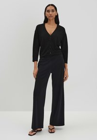 Schwarzer gerippter Cardigan mit V-Ausschnitt und Knopfverschlüssen, kombiniert mit schwarzen weiten Hosen und flachen Sandalen, auf einem neutralen Hintergrund präsentiert.