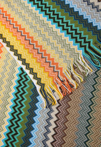 Capa com padrão de zig-zag multicolorido e bordas franjas; apresenta tons de laranja, azul, verde e castanho; feita de tecido texturizado.