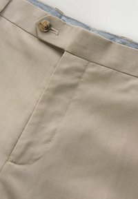 Pantalon beige à la texture lisse, doté d'une fermeture à un bouton et d'un détail de poche discret. L'intérieur présente un motif en chevrons bleu.