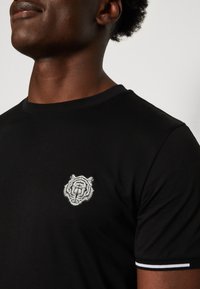 Camiseta de manga corta negra con un emblema de tigre bordado en blanco en el pecho, con acentos en blanco contrastantes en los dobladillos de las mangas.