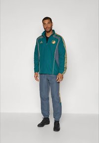 Mees seisab, seljas tealkolline tõmbluku ja kuldsete triipudega spordijakk, millele sobivad hallid treeningpüksid kuldsete triipudega ja mustad jooksujalatsid.